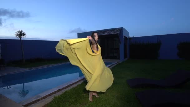 Linda mulher de moda com cabelo escuro em vestido amarelo elegante posando e dançando ao lado da piscina na luxuosa villa - vídeo em câmera lenta — Vídeo de Stock