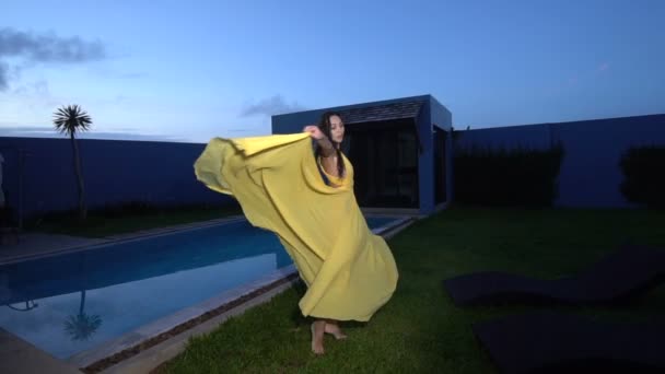 Magnifique femme de mode aux cheveux foncés dans une élégante robe jaune posant et dansant au bord de la piscine à la villa luxueuse - vidéo au ralenti — Video