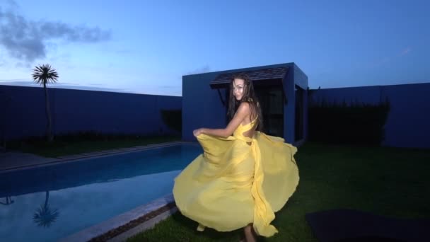 Preciosa mujer de moda con cabello oscuro en elegante vestido amarillo sonriendo y bailando junto a la piscina en la lujosa villa - video en cámara lenta — Vídeos de Stock