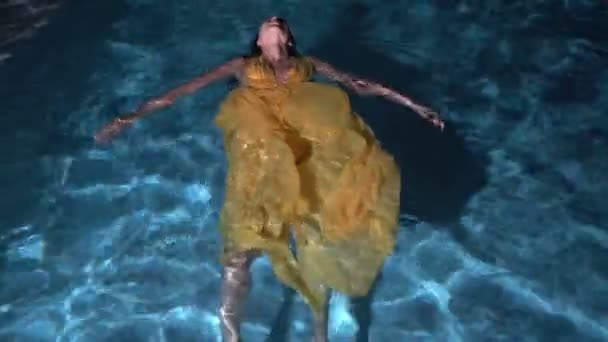 Κορυφαία θέα της πανέμορφης μόδας γυναίκα με σκούρα μαλλιά σε κομψό κίτρινο φόρεμα που βρίσκεται στο νερό της εξωτερικής πισίνας τη νύχτα — Αρχείο Βίντεο