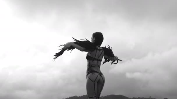 Back view of beautiful seductive angel woman wearing lingerie and leather belts standing on the roof with wind in her wings over cloudy sky - black and white video — Stock Video