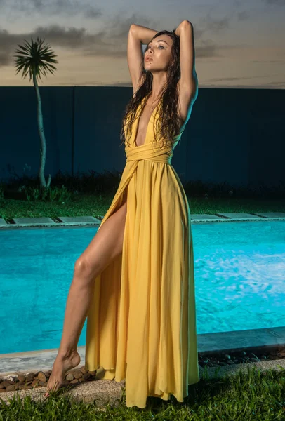 华丽的时尚黑发女人在优雅的黄色礼服摆姿势在游泳池旁边的游泳池在豪华别墅在日落背景的晚上 — 图库照片
