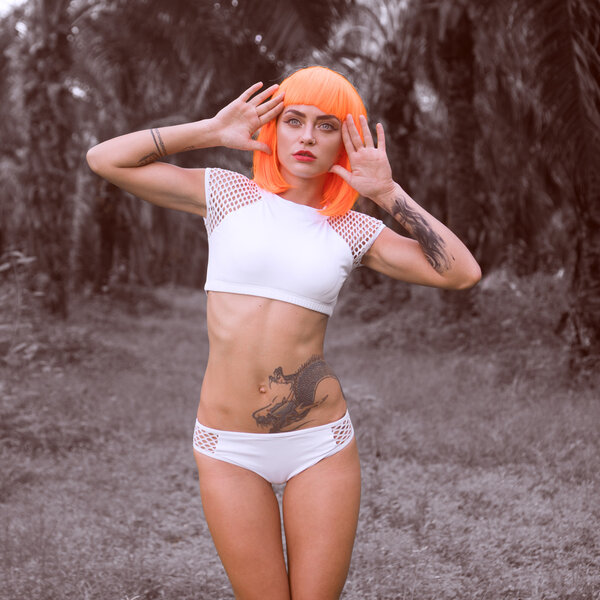Sexy beautiful woman in modern futuristic style posing in palm trees forest. Creative look of tattooed woman wearing white bikini and orange wig