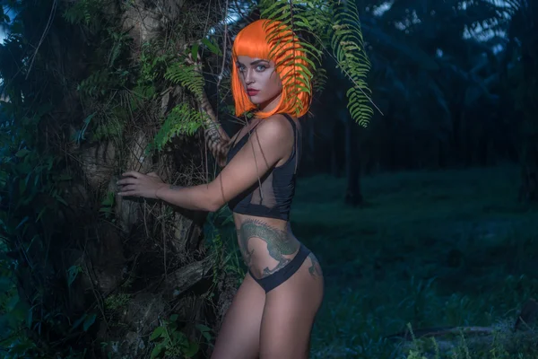 Sexy mooie vrouw in moderne futuristische stijl poseren in palmbomen bos. Creatieve look van getatoeëerde vrouw met zwarte Romper en oranje pruik op zoek naar de camera — Stockfoto