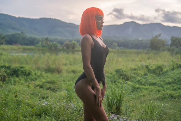 Mulher bonita sexy em estilo futurista moderno posando no campo verde sobre fundo incrível céu pôr do sol. Look criativo de mulher tatuada vestindo bodysuit preto e peruca laranja — Fotografia de Stock