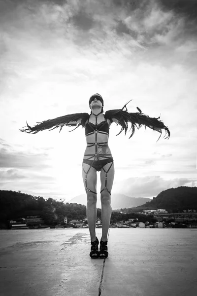 Schwarz-Weiß-Foto der schönen verführerischen Engelsfrau in Dessous und Ledergürteln, die mit Wind auf den Flügeln über dem bewölkten Himmel auf dem Dach steht — Stockfoto