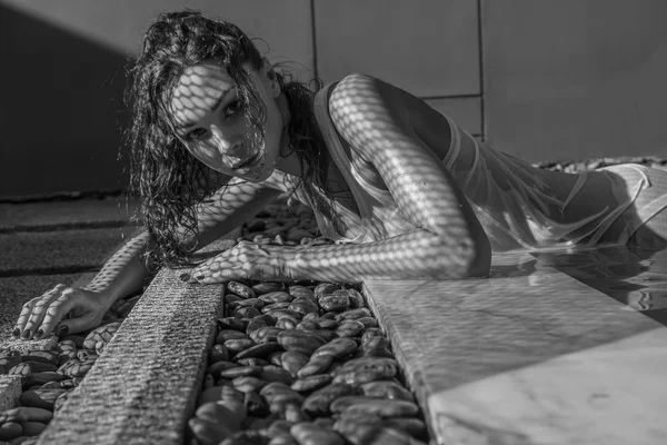 Foto de cerca en blanco y negro de una hermosa morena seductora posando en la piscina al aire libre durante el soleado día de verano con reflejo de la red de sombras en su rostro y cuerpo — Foto de Stock