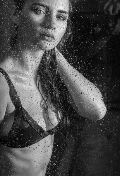 Black and white photo of beautiful young brunette woman wearing black underwear looking through the window during rainy day