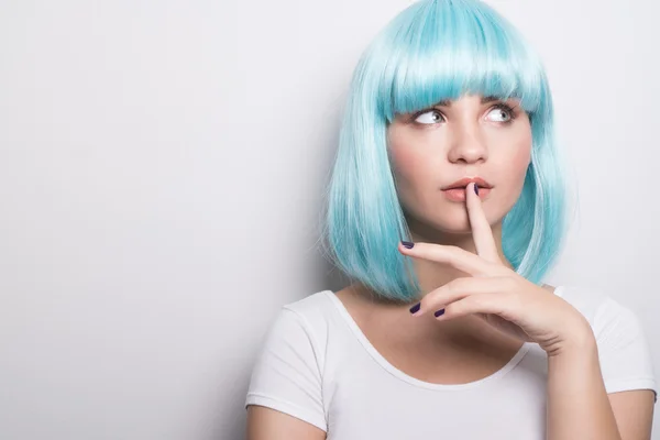 Nahaufnahme Porträt eines frechen jungen Mädchens im modernen futuristischen Stil mit blauer Perücke mit neugieriger Miene und Finger auf der Lippe beim seitlichen Blick über weißen Wandhintergrund mit Kopierraum — Stockfoto