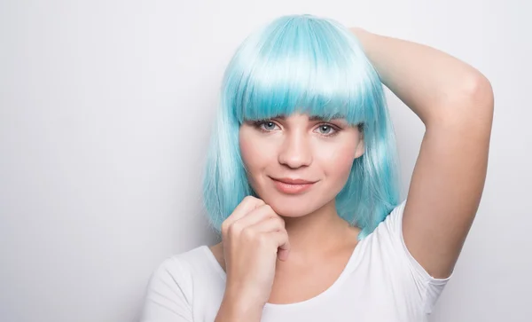 Jeune fille insolente dans un style futuriste moderne avec une perruque bleue souriante et regardant dans la caméra avec ses mains sur la tête sur fond de mur blanc avec copyspace — Photo