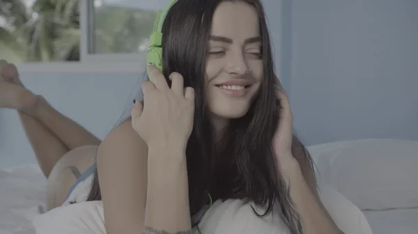 Belle jeune femme nue brune souriante tout en écoutant de la musique à l'aide de son smartphone et de écouteurs verts allongés sur le lit dans la chaude matinée d'été — Photo