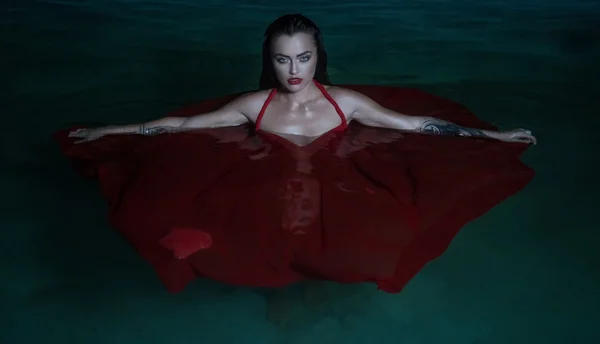 Beautiful mysterious woman looking like a vampire wearing red dress looking into the camera ehile standing in the pool at night — Stock Photo, Image