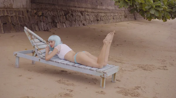 Sexy mooie vrouw in moderne futuristische stijl poseren op de beschadigde houten blauwe zonnebank. Creatieve look van vrouw op het strand dragen bikini en blauwe pruik — Stockfoto