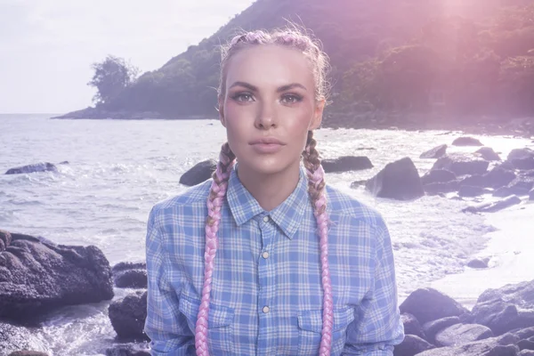 Ritratto di giovane graziosa ragazza con trecce rosa che indossa camicia a quadri blu guardando nella macchina fotografica mentre seduta sulla roccia durante la giornata estiva sul mare e sullo sfondo del cielo — Foto Stock