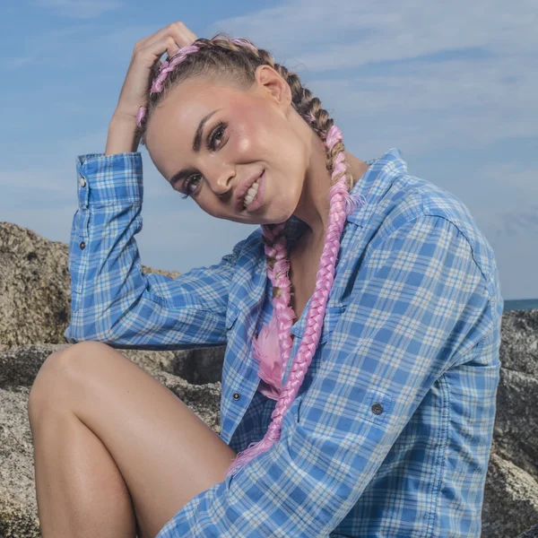Young pretty girl with pink plaits wearing blue checked shirt smiling and looking into the camera while sitting on the rock during summer day over sky background — Stock Photo, Image