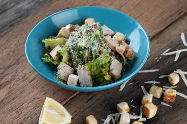 Tavuklu Cesar salatası, kıtır ekmek, rendelenmiş peynir ve limon ahşap kırsal masada izole edilmiş.