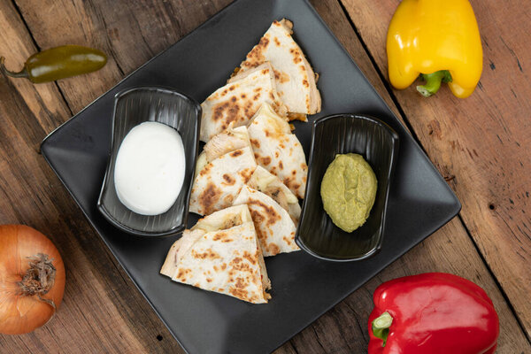 A toasted tortilla with chicken, peppers, red onion and mozzarella served with a guacamole dip and sour cream isolated on wooden table