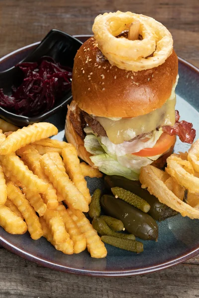 Big burger with beef, bacon, tomato, cheese, lettuce and onion served with french fries, onion rings, pickles and beetroot salad on the side isolated on rustic wooden table