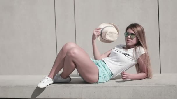 Chica con el pelo largo con gafas de sol y sombrero, día caluroso de verano, posando contra la pared, urbano — Vídeo de stock