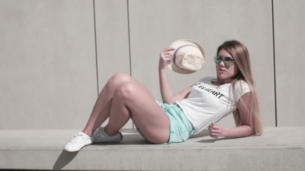 Mädchen mit langen Haaren mit Sonnenbrille und Hut, sommerlich heißer Tag, posiert gegen die Wand, urban