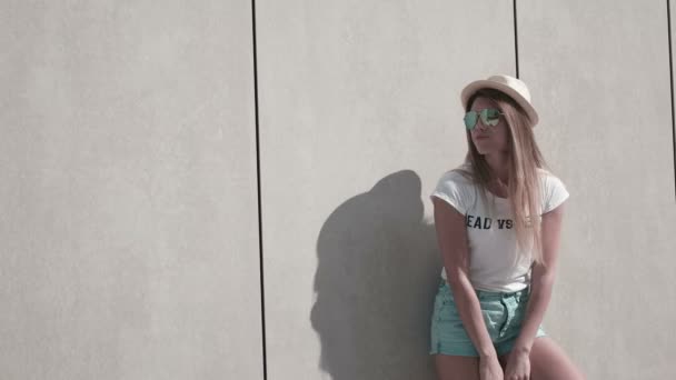 Chica con el pelo largo con gafas de sol y sombrero, día caluroso de verano, posando contra la pared, urbano — Vídeo de stock