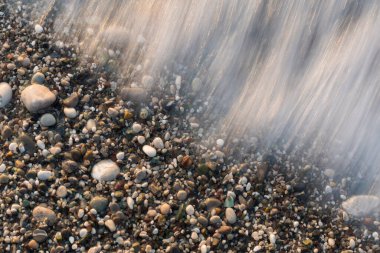 Yüksek açılı, uzun pozlu bir çekim, çeşitli pürüzsüz nehir taşları ve çakıl taşlarının üzerinden geri akan bir deniz dalgasının hareketini yakalar. Su, renkli kayaların topraklanmış dokusuna karşı yumuşak, ipeksi beyaz izler gibi görünür. Doğal ışık.