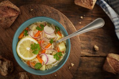 Lezzetli ve besleyici bir kase tavuk çorbası taze limon dilimleri, havuç ve soğanla servis edilir. Tabak, kırsal bir ahşap masada, üzerinde tam tahıllı ekmek parçaları bulunan parlak mavi bir seramik kasede sunuluyor. Bu kaliteli yemek fotoğrafçısı şapkası.
