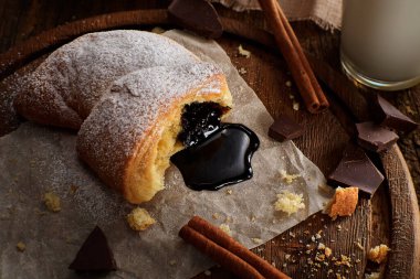 Parşömen kağıdına sızan koyu çikolata soslu sıcak, pudralı bir kruvasanın enfes yakın çekimi. Sahne, çikolata parçacıkları, tarçın çubukları ve bir bardak sütle kırsal bir tahta üzerinde güzelce düzenlenmiş.