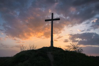 Uzun bir Hristiyan haçının güçlü silueti çimenli bir tepenin üzerinde duruyor dramatik bir günbatımı gökyüzüne karşı altın bulutlarla, inancı, umudu ve maneviyatı simgeliyor..