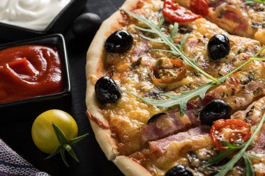 Yakın plan, kıtır jambon, siyah zeytin, vişneli domates, erimiş peynir ve taze roka yapraklı taze gurme pizzası. Koyu ahşap arka planda servis edilir. Yanında domates sosu ve beyaz krema kâseleriyle..