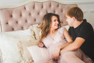 Genç hamile kız, yakışıklı bir adamla rahat bir yatakta uzanıyor gülümsüyor ve sarılıyor.