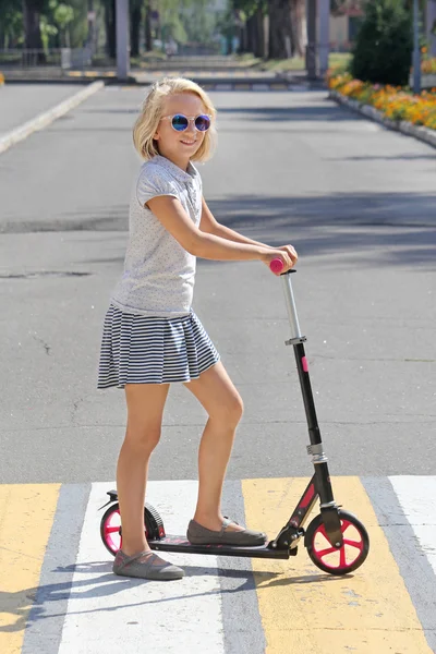 Mädchen im Schulalter auf einem Motorroller. überquert die Straße auf einem Fußgängerüberweg. Stockbild