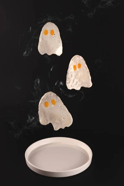 Жареные яйца в виде призраков летают над белой тарелкой на черном фоне — стоковое фото