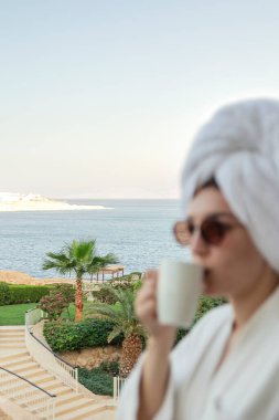 Gözlüklü, bornozlu ve havluyla kahve içen kadının bulanık görüntüsü.