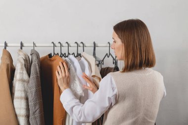 Kadın milenyum stilisti giyinme odasındaki şık korkuluklardan kıyafet seçiyor.