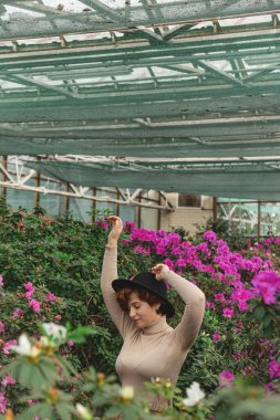 Seranın yeşil bitkilerinin arasında gülümseyen şapkalı, iri yarı, güzel bir kız.