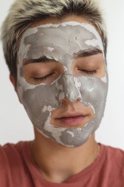 Kapalı gözlü bir kadının görüntüsü yüzüne gri yeşil kil maske takıyor. Günlük güzellik derisi bakım rutini. Görüntüyü kapat. 