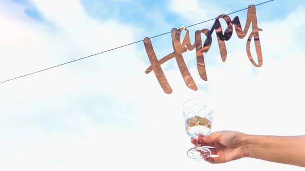 Goldenes Wort glücklich hängt an Girlande vor blauem Himmel mit Wolken neben Frauenhand mit Glas Wein. — Stockfoto