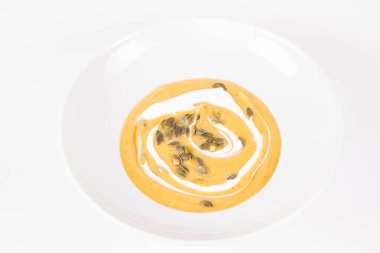 Beyaz arka planda bir tabakta krema ve kabak tohumlarıyla süslenmiş balkabağı çorbası.