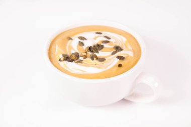 Beyaz arka planda bir kasede krema ve kabak tohumlarıyla süslenmiş balkabağı çorbası.