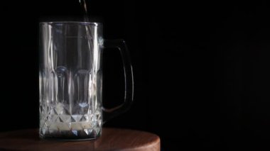 Siyah arka planda, hafif köpüklü bir bira, ahşap bir stantta yarım litre bardağa dökülür. Bira kenardan dökülüyor..