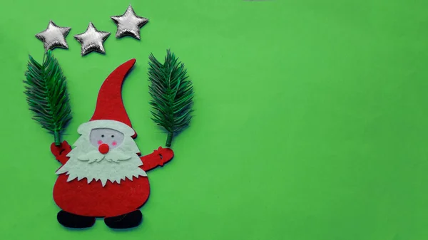 El yapımı Noel kompozisyonu. Elinde köknar dalları tutan Noel Baba 'nın bir resmi var, başının üstünde yeşil bir zemin üzerinde keçe ve kontrplak parçalarından yapılmış üç gümüş yıldız var. Resmin sağında boş alan.