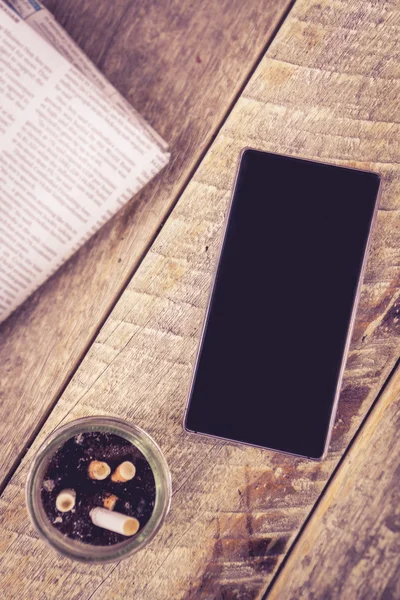 Teléfono móvil cenicero periódico en mesa de madera —  Fotos de Stock