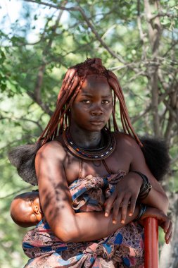 NAMIBIA, OMUSATI REGION, 7 MAYIS: geleneksel saç stili ve sırtında uyuyan bebek Himba kadın portresi. Himba, Namibya 'nın kuzeyindeki Namibya' nın yerlilerindendir.