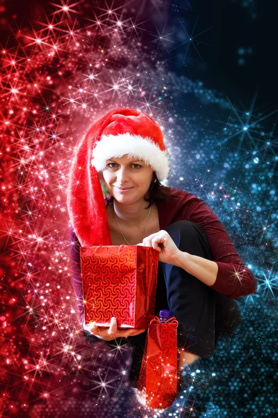 Schöne Frau mittleren Alters in rotem Party-Weihnachtsmann-Hut sitzend mit Geschenken, mit abstraktem Hintergrund — Stockfoto