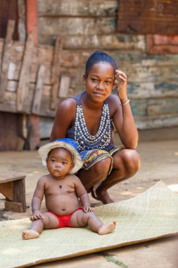 Brickaville, Toamasina Eyaleti, Madagaskar - 17 Ekim 2016: Toamasina bölgesindeki ahşap bir evin dışında örülmüş bir paspasın üzerinde oturan güneş şapkası takan bir kadın ve bir bebeğin portresi.