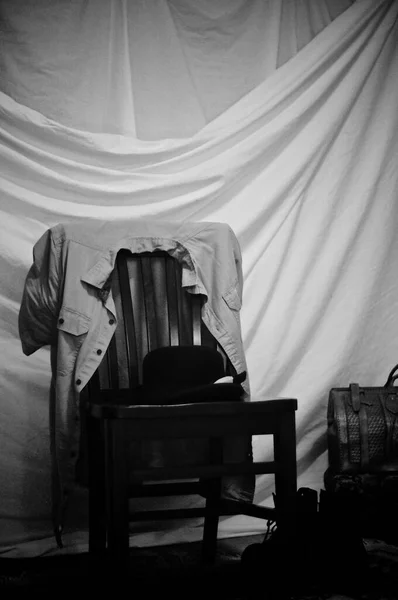 A black and white back stage still life with draped curtain backdrop, leather bag, boots, and a man\'s hat and shirt on wooden chair.