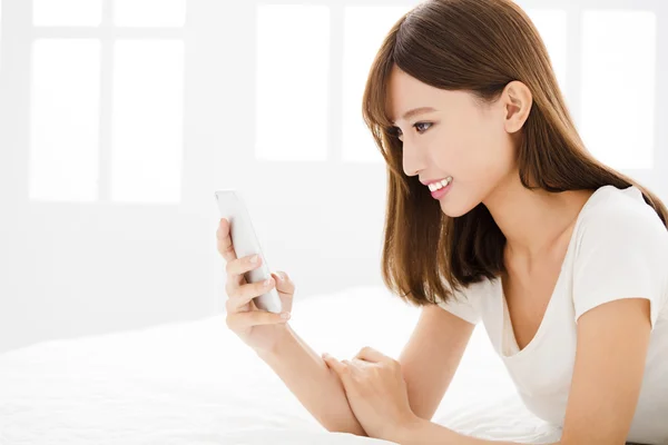 Unga leende kvinna läser smart telefon på sängen — Stockfoto