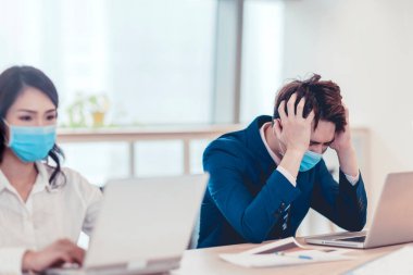Stresli iş adamı yüz maskesi takıyor ve ofiste dizüstü bilgisayarla çalışıyor.