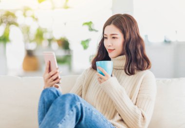 Kanepede oturan bir fincan kahve, çay ve akıllı telefon izleyen bir kız. 